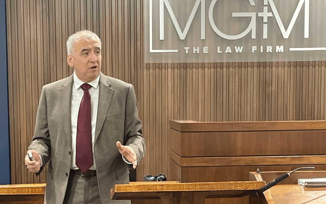 A man wearing a brown suit stands in front of a sign for a law firm